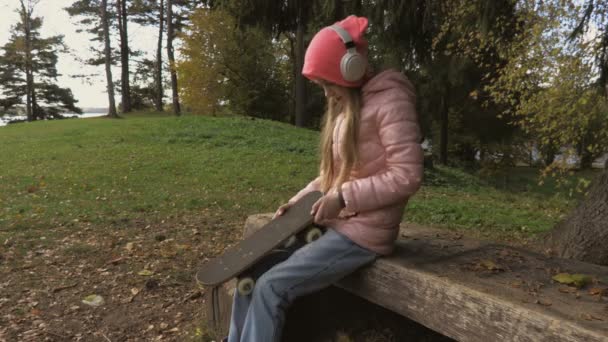 ヘッドフォンを使用して ベンチでスケートボードを保持する女の子 — ストック動画