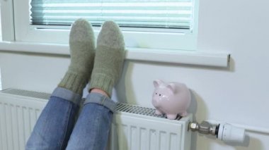 Kışın radyatörde domuz kumbarası yakınında ayaklarını ısıtan bir kadın.