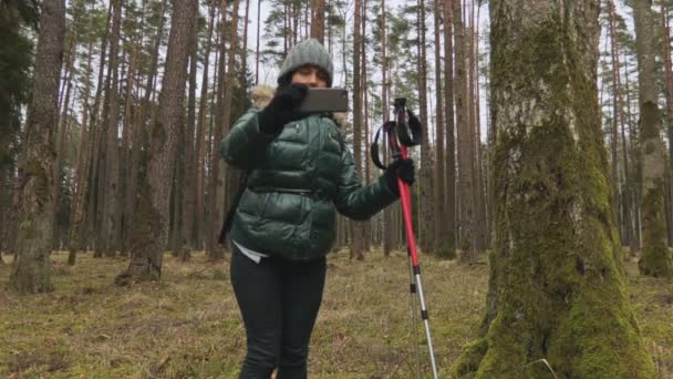 スマホを使ってバックパックやトレッキングスティックを持つ森の中の女性観光客 — ストック動画