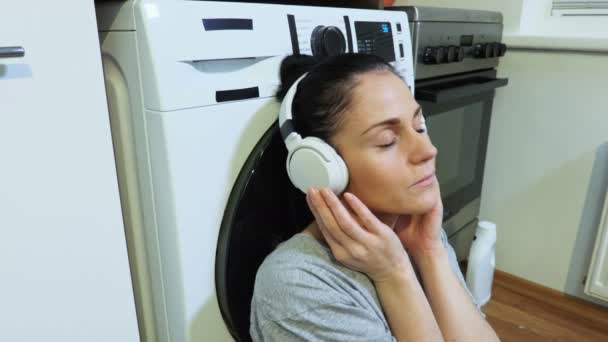 坐在洗衣机旁边 用耳机听音乐的女人 — 图库视频影像