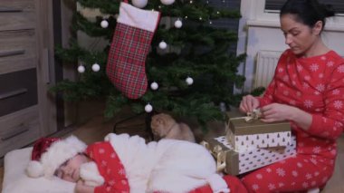 Noel pijamalı bir kadın Noel ağacında uyuyan küçük bir kızın yanında hediye kutuları düzenliyor.