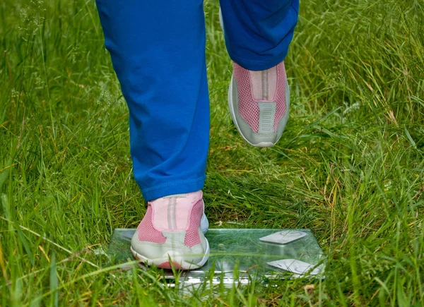Sport sportswoman fat woman lose weight is standing on the scales legs right leg raised blue sports trousers knee-deep in pink sneakers glass transparent scales on green grass blurred background front view