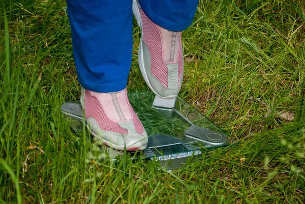 Sport athlete thick woman lose weight standing left sideways on the scales legs right leg raised blue sports trousers knee-deep in pink sneakers glass transparent scales side view on green grass blurred background