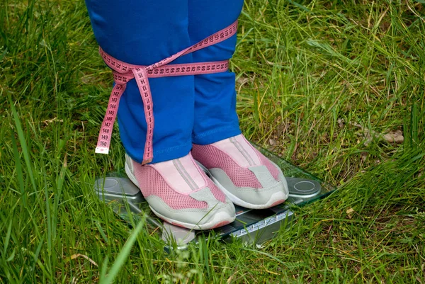 Sport sportswoman fat woman lose weight is standing on the scales legs connected pink measuring tape, figures, blue sports trousers knee-deep in pink sneakers glass transparent scales on green grass blurred background