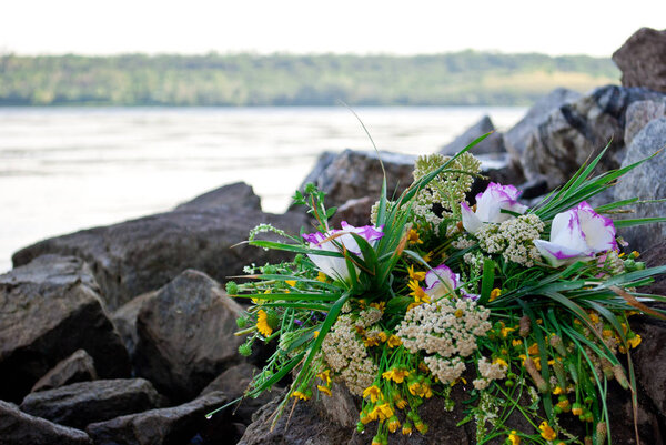 One beautiful large bouquet of a wreath white purple yellow beautiful fresh wildflowers and green grass lies on rocks against the background of the Ukrainian river Dnieper on the feast of Ivan Kupala