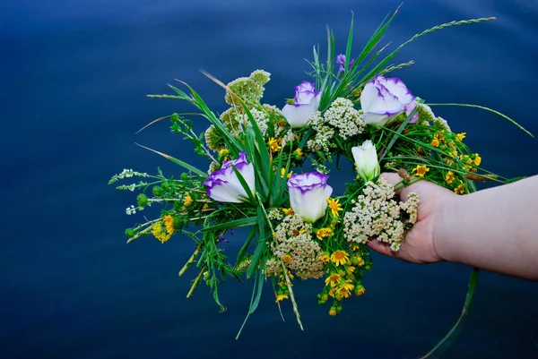 One big beautiful bouquet of a wreath of wildflowers with yellow white and purple flowers and green grass is held by a female hand girl over a river in the evening holiday of Ivan Kupala