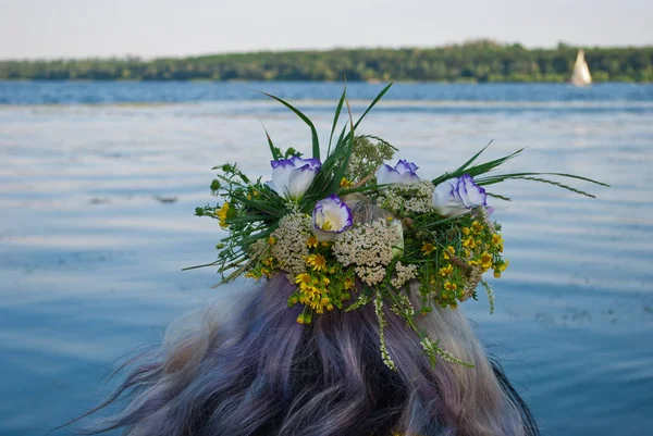 One big beautiful bouquet wreath of wild flowers with yellow white and purple flowers and green grass on the head of the girl hair blonde over the water river in the evening holiday of Ivan Kupala