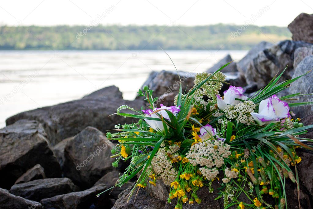 One beautiful large bouquet of a wreath white purple yellow beautiful fresh wildflowers and green grass lies on rocks against the background of the Ukrainian river Dnieper on the feast of Ivan Kupala