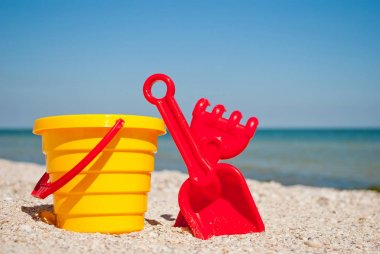 Sarı çocuk kırmızı kulplu plastik kırmızı kürek kemiği kova ve komisyon sol tarafta bir mavi deniz arka plan ve mavi gökyüzü sarı kum plaj deniz shore deniz kabuklarını yaz tatili güneşli yaz günü, parlak güneş