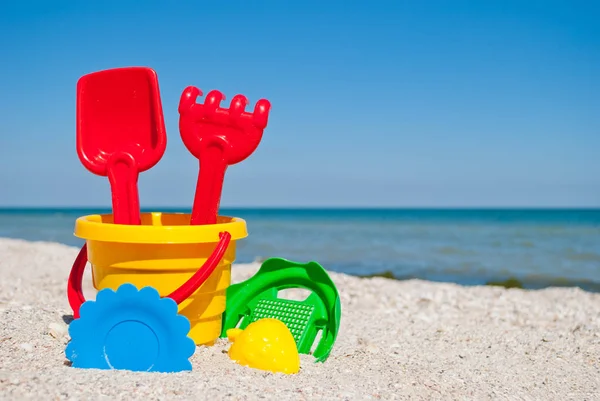 Sarı bebek kova kırmızı kolu, kırmızı plastik spatula ve komisyon, yeşil elek, çilek, mavi tabak, mavi deniz ve gökyüzü, sarı kum plaj deniz kabuklarını deniz yaz tatili güneşli sarı kum formu ile