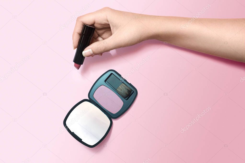 Female hand holding lipstick and blusher with mirror on pink background