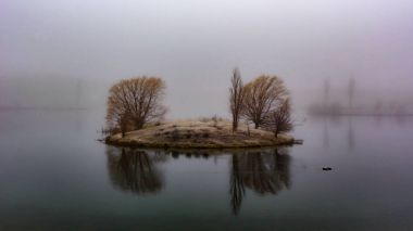 View of a small land in the middle of the lake during a foggy da clipart