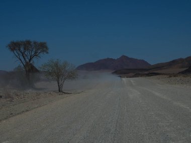 Açık mavi gökyüzü ile çöl ve dağ manzara arasında tozlu asfaltsız yol ile offroad araç sürüş