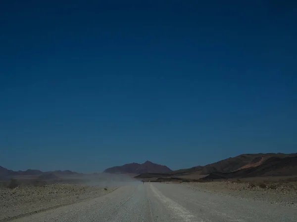 Viaje por carretera a través de un polvoriento camino sin pavimentar entre el desierto y el paisaje de montaña con cielo azul claro —  Fotos de Stock