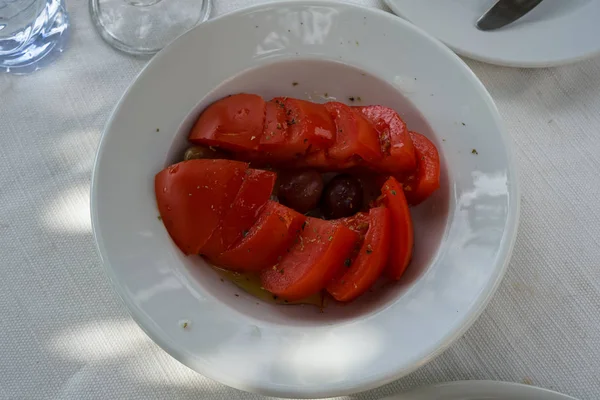 Fresh mediterranean tomato salad with olive fruit, oil and oregano in white round dish serving on street side cafe table on sunshine day