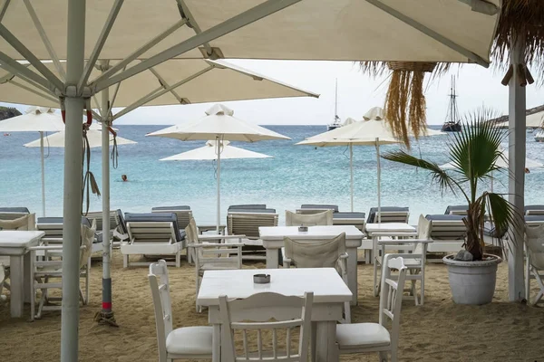 Restaurant tables and chairs setup under umbrella in white color and long chair in blue on Ornos sand beach with seaview and sailing boat view