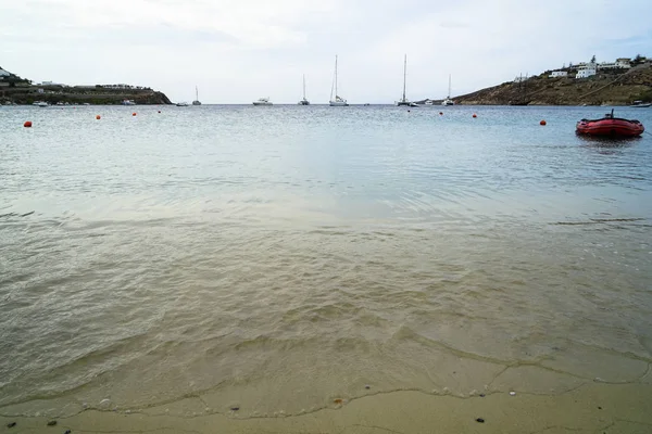 Morbido paesaggio bellissimo paesaggio marino e spiaggia di sabbia naturale con barche a vela, yacht, gommone rosso, cielo limpido, montagna e sfondo bianco edificio, spiaggia di Ornos — Foto Stock