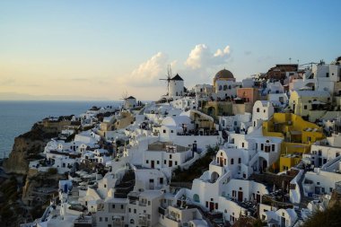 Işık Oia yel değirmeni ve beyaz bina townscape uçsuz bucaksız Okyanusu yumuşak bulut ve açık mavi gökyüzü arka plan ile karşı karşıya ada dağ boyunca akşam içinde panoramik sahne