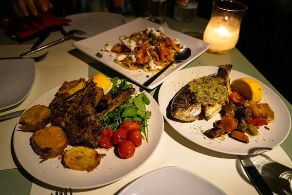 Delicious candle light dinner table serving fresh grilled lamb and whole sea bream fish with roasted potato, tomato, carrot, pepper, mushroom, zucchini, rocket salad and yellow lime; and seafood spaghetti pasta on white plate