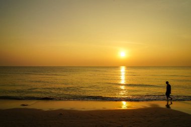 Deniz manzarası ile sabah gündoğumu sahnesinde kum plajda yalınayak yürüyen bir adam siluet, dalga su yansıması, yumuşak geniş turuncu renk gökyüzü arka plan tonları