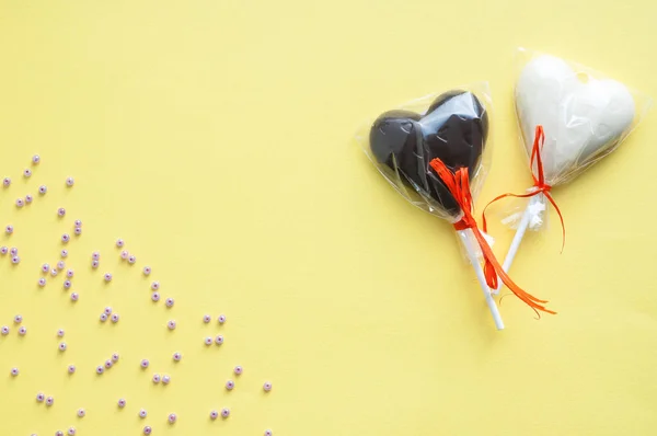 Dulces de chocolate blanco y negro en forma de corazón en un palo, vendados con una cinta rosa y muchas cuentas de color lila sobre un fondo amarillo claro. Tarjeta horizontal para el Día de San Valentín . — Foto de Stock