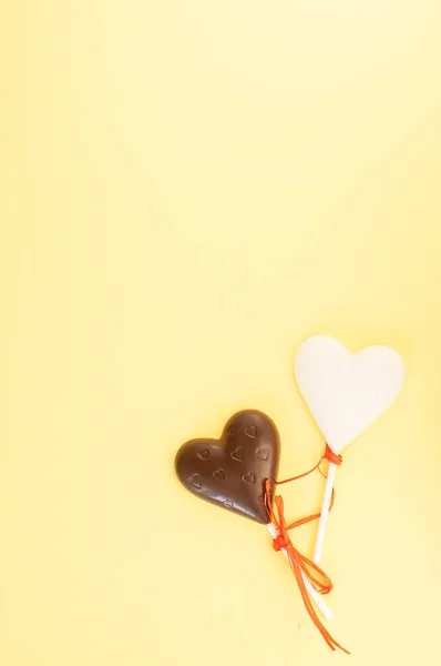 Dulces de chocolate blanco y negro en forma de corazón sobre un palo, vendados con una cinta naranja sobre un fondo amarillo claro. Tarjeta vertical para el día de San Valentín . — Foto de Stock