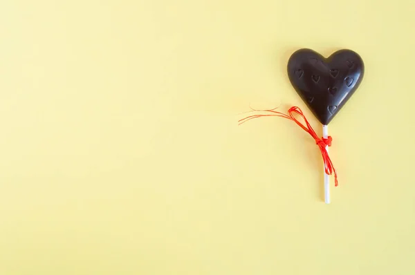 Un caramelo de chocolate negro en forma de corazón en un palo, vendado con una cinta naranja sobre un fondo amarillo claro. Tarjeta para el Día de San Valentín . — Foto de Stock
