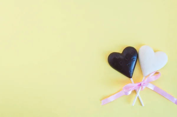 Dulces de chocolate blanco y negro en forma de corazón en un palo, vendados con una cinta rosa sobre un fondo amarillo claro. Tarjeta para el Día de San Valentín . — Foto de Stock