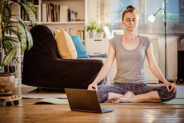 Evde yoga yapan bir kadın dizüstü bilgisayardan video izliyor.