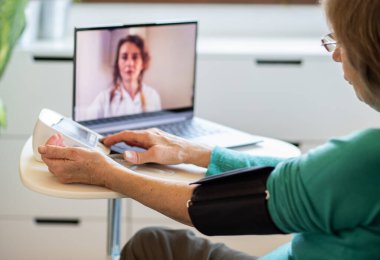 Teletıp konsepti. Yaşlı bir kadın doktoruyla internetten konuşuyor ve tansiyonunu ölçüyor.
