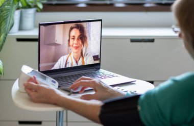 Teletıp konsepti. Yaşlı bir kadın doktoruyla internetten konuşuyor ve tansiyonunu ölçüyor.