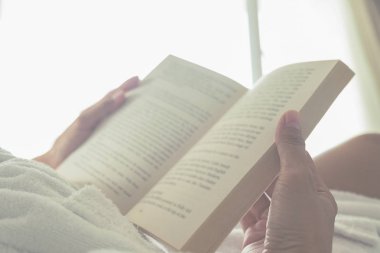 Bir fincan kahve ile yatağın üstüne oturan ve bir kitap okuma güzel Asyalı kadın. Bir kitap ve kahve ile sabah. Rahatlatıcı kavramı. Retro filtre efekti, yumuşak odak, seçici odak