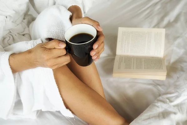 Красивая азиатская женщина сидит на кровати с чашкой кофе и читает книгу. Утро с книгой и чашкой кофе. Расслабляющая концепция. Эффект ретро-фильтра, мягкая фокусировка, избирательная фокусировка — стоковое фото