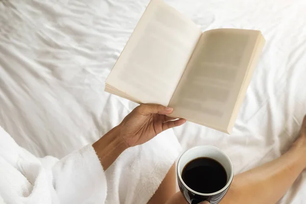 Belle asiatique femelle assise sur le lit avec une tasse de café et la lecture d'un livre. Bonjour avec un livre et une tasse de café. Concept relaxant. Effet filtre rétro, mise au point douce, mise au point sélective — Photo