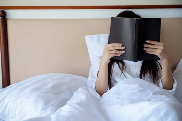 Hermosa mano asiática mujer sosteniendo una taza de café en la cama, leyendo un libro y utilizando la computadora con luz de la mañana. Concepto relajante. Efecto filtro retro, enfoque suave, enfoque selectivo . — Foto de Stock