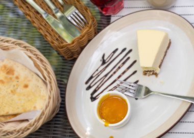 Büyük beyaz çanak, mango soslu krem peynirli kek. Parlak bokeh arka plan. Çatal bıçak takımı, beyaz peçeteler, kahve fincanı, şekerlik, hasır sandalyeleri sepetle. Tatlı. Vişne suyu