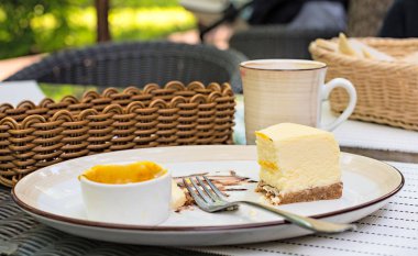 Büyük beyaz çanak, mango soslu krem peynirli kek. Parlak bokeh arka plan. Çatal bıçak takımı, beyaz peçeteler, kahve fincanı, şekerlik, hasır sandalyeleri sepetle. Tatlı. Vişne suyu