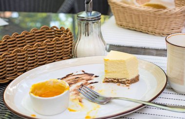 Büyük beyaz çanak, mango soslu krem peynirli kek. Parlak bokeh arka plan. Çatal bıçak takımı, beyaz peçeteler, kahve fincanı, şekerlik, hasır sandalyeleri sepetle. Tatlı. Vişne suyu