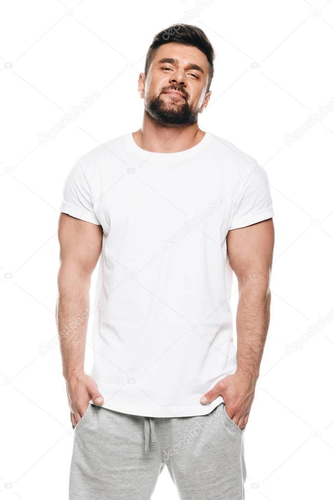 Portrait of muscular bearded man standing with hands in pockets and looking at camera isolated on white