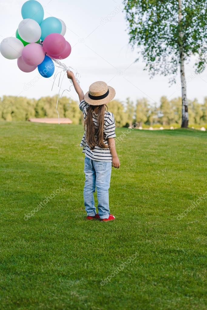 Back view of cute little girl in straw hat holding colorful balloons in park