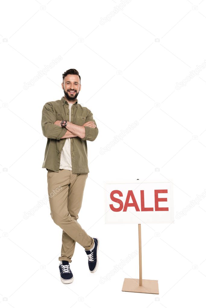 Smiling man with arms crossed standing near sale sign isolated on white