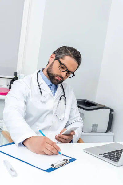 Doctor using smartphone and writing in clipboard — Stock Photo, Image