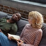 Africano americano hombre escuchar música mientras caucásico novia lectura libro en sofá en casa