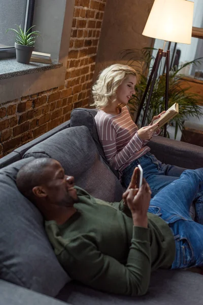 非洲裔美国人使用智能手机 而白种人的女朋友在家里的沙发阅读书 — 免费的图库照片