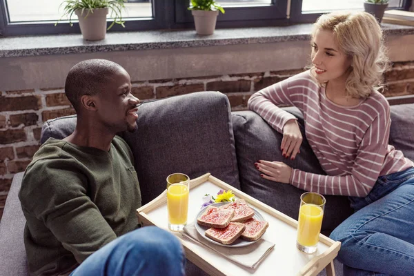 一緒に家で朝食をとりながらソファーに座っていた多文化の若いカップルの肖像画  — 無料ストックフォト