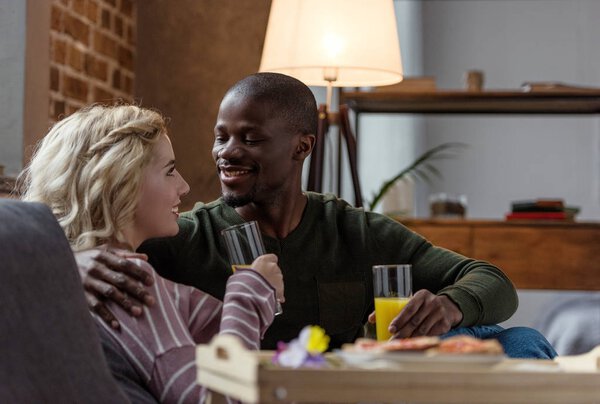 side view of smiling interracial couple with glasses of juice looking at each other at home