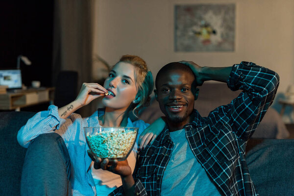 portrait of multiethnic couple eating popcorn and watching film together on sofa at home