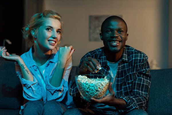 portrait of cheerful multiethnic couple watching tv together on sofa at home