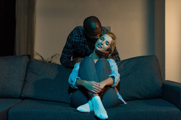Африканский американец целует улыбающуюся кавказскую девушку на диване дома
