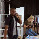 Afikanisch-amerikanischer Fotograf spricht auf Smartphone, während kaukasischer Kollege Fotos im Studio auswählt